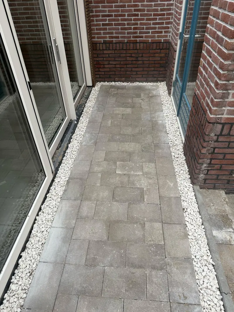 Pad van grijze betontegels met een rand van wit grind langs schuifpuien